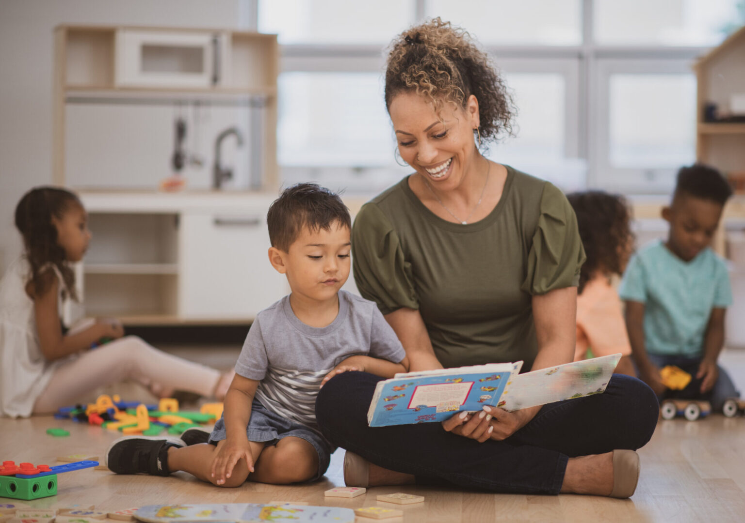 A preschool boy of Hispanic ethnicity sits next to his teacher as she is reading him a book on the classroom floor.